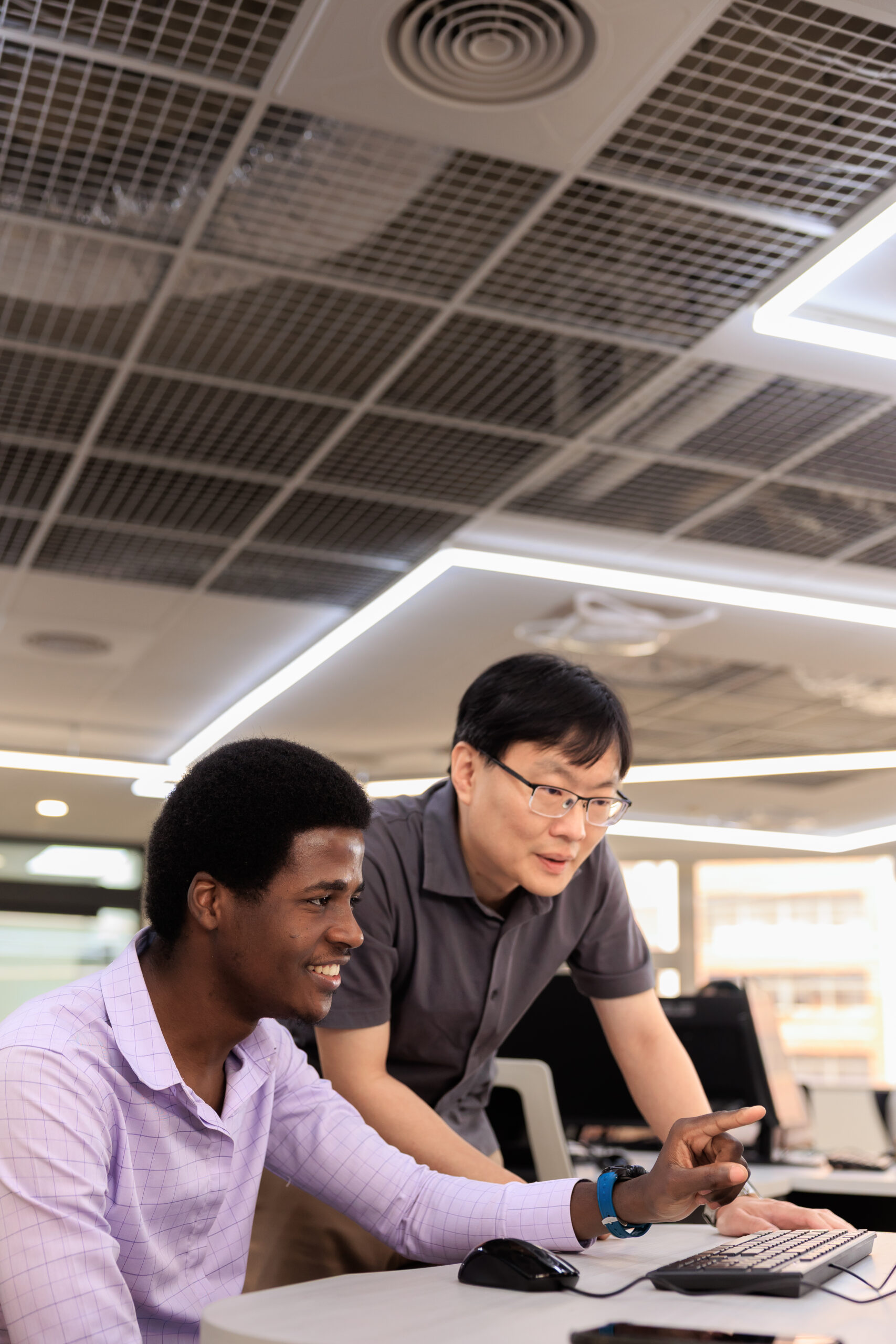 Adult Black male international student and East Asian male professor collaborating at a workstation in a modern computer lab at I-Shou University, Taiwan. The professor points and mentors the student